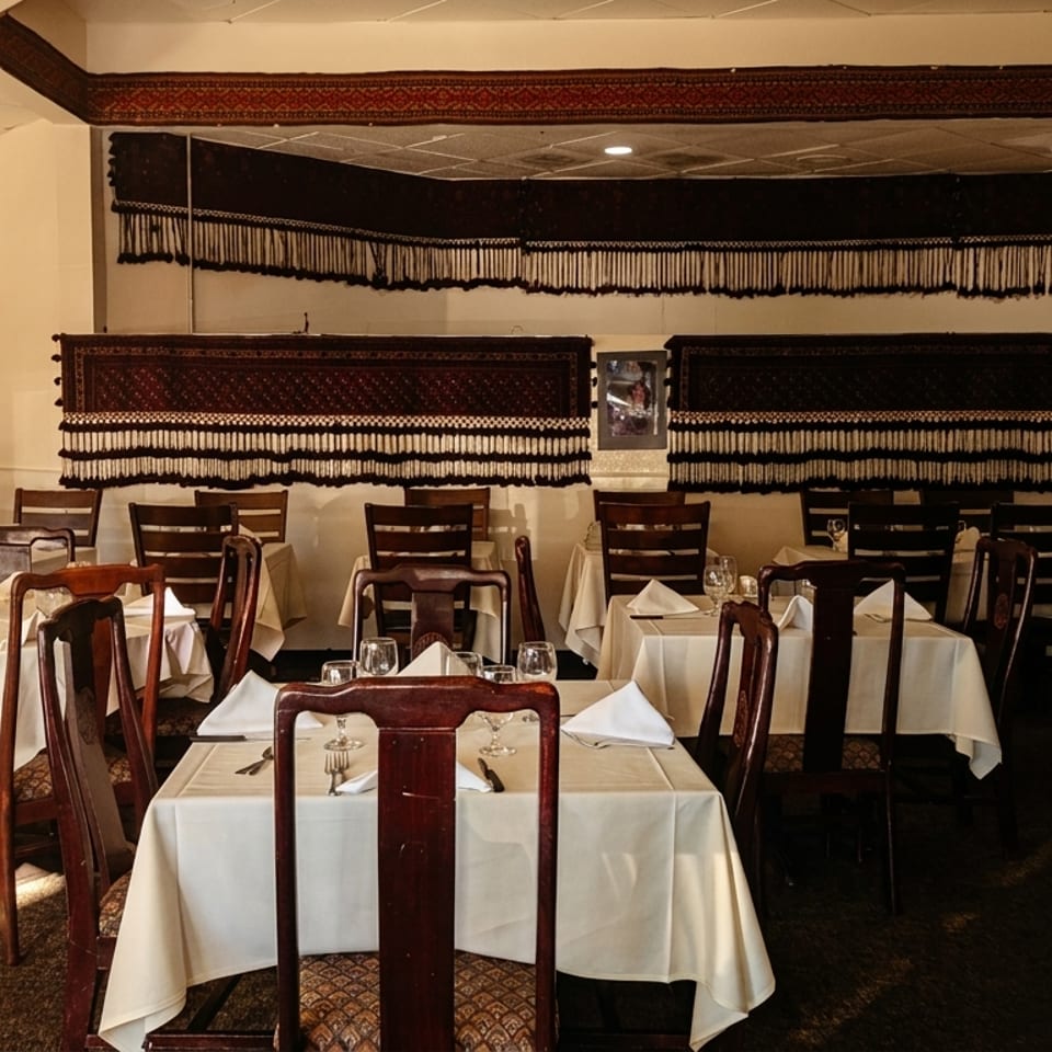 Empty restaurant dining room with white tablecloths, dark wooden chairs, and decorative tapestries on the walls.