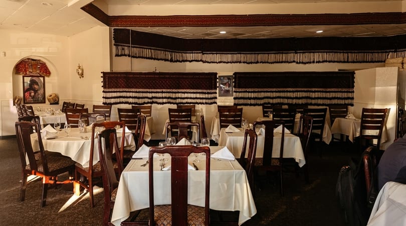Empty restaurant dining room with white tablecloths, dark wooden chairs, and decorative tapestries on the walls.