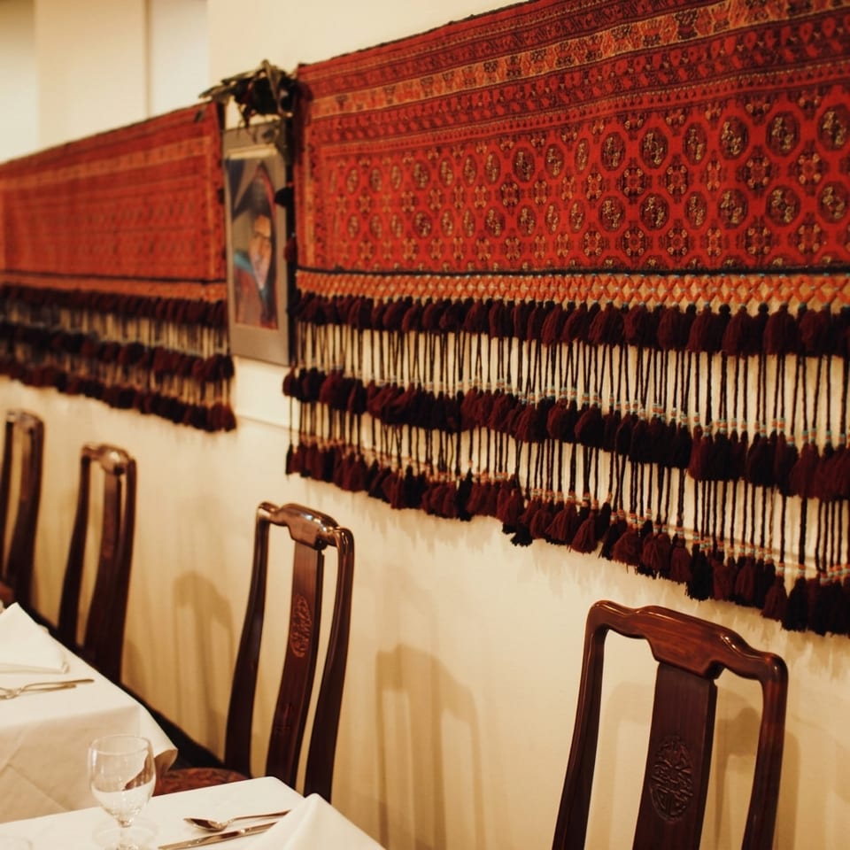 Restaurant interior with white tablecloths, dark wooden chairs, and a red patterned tapestry with tassels on the wall.