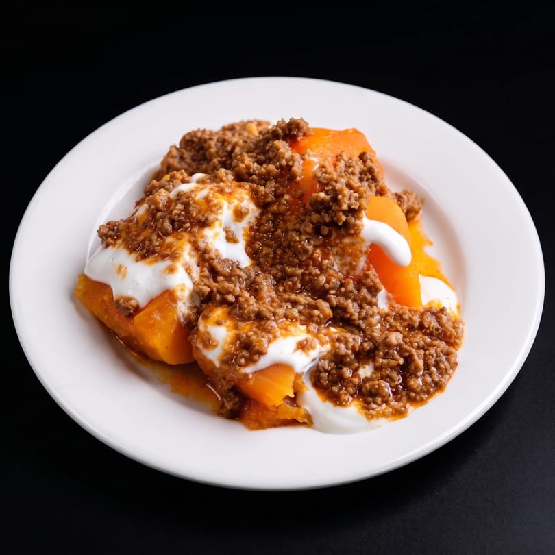 A white plate holds a dish of orange sweet potatoes topped with ground meat and white sauce.
