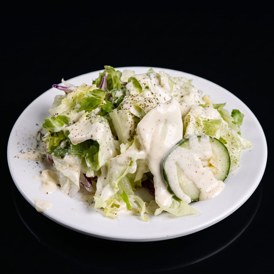 Fresh green salad with creamy dressing, black pepper, and cucumber slices on a white plate.