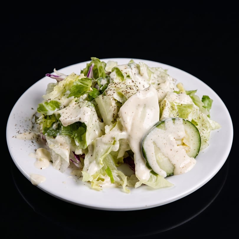 Fresh green salad with creamy dressing, black pepper, and cucumber slices on a white plate.
