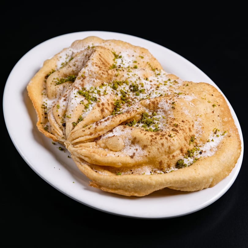 Golden fried pastries dusted with powdered sugar, cinnamon, and chopped pistachios on a white plate.